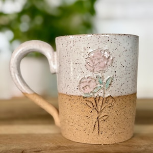 May include: A handmade ceramic mug with a speckled white upper half and a tan lower half. The mug features a floral design with pink flowers and green leaves. The handle is white with a tan base.