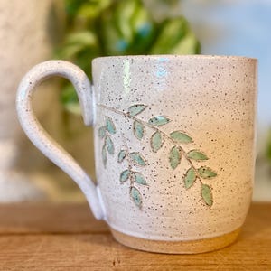 May include: A ceramic mug with a speckled off-white glaze and a curved handle. The exterior features a hand-carved design of two leafy branches with light green glaze filling the leaves. The base of the mug is unglazed, revealing a tan clay color.