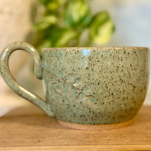 May include: A ceramic mug with a speckled light green glaze and a glossy finish. The mug features a curved handle and an embossed leaf pattern on the side. The base of the mug is unglazed, showing the natural tan color of the clay material.