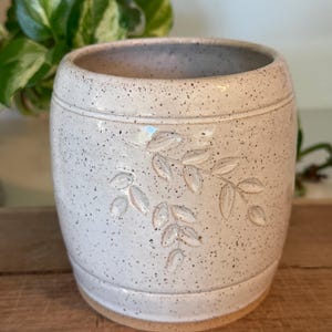 May include: A cylindrical ceramic planter with a speckled off-white glaze and a debossed leaf pattern on the front. The vessel features a smooth, rounded rim and a plain, unglazed base, showcasing a natural clay texture at the bottom edge.