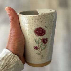May include: A speckled off-white ceramic tumbler featuring a raised, textured design of three red roses with green stems. The cup has a smooth, tapered cylindrical shape with a distinct notch at the rim and a plain, unglazed tan base at the bottom.