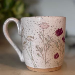 May include: A ceramic mug with a speckled off-white glaze featuring embossed floral designs in shades of pink and deep red. The mug has a curved handle on the left side and a natural, unglazed tan clay base at the bottom, showcasing a textured finish.