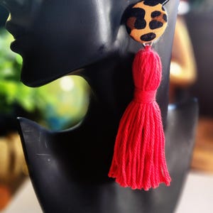 May include: A single earring featuring a leopard print button and a long, bright red tassel. The earring is displayed on a black mannequin head. The background is blurred, with hints of green and gold.