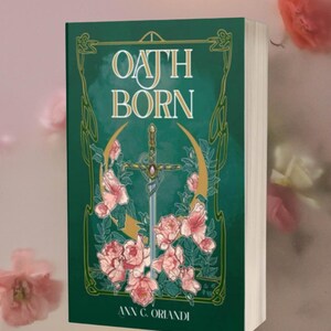 May include: A hardcover book titled Oath Born by Ann C. Orlandi with a deep green cover featuring a central sword illustration, a golden crescent moon, and pink floral accents framed by decorative gold lines and intricate border patterns.