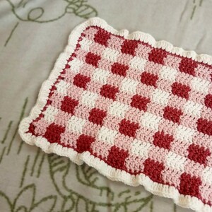 Peut inclure: Carré crocheté fait main avec un motif à carreaux rouge, blanc et rose. Les bords sont finis avec une bordure blanche festonnée. Il pourrait s'agir d'un dessous de verre ou d'une petite pièce décorative.