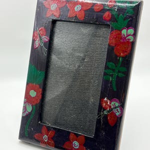 May include: A rectangular picture frame with a dark purple base color, featuring hand-painted red flowers with light blue centers and green leaves. The frame includes two small, detailed butterfly illustrations in pink and green on the sides.