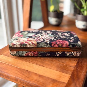 May include: A rectangular clutch bag featuring a dark fabric exterior with a multi-colored floral pattern in shades of red, pink, white, and green. The bag has a metallic gold-toned frame that runs along the center edge, providing a structured closure.