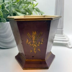 May include: A hexagonal planter with a deep maroon finish and gold-toned trim along the top and base edges. The front panel features a gold-coloured graphic illustration of a flaming torch crossed with two arrows, accented by a decorative ribbon.
