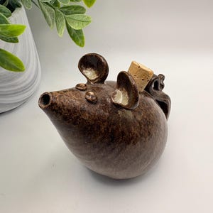 May include: A dark brown ceramic pourer shaped like a mouse with a glossy finish, two rounded ears, a pointed spout, and a small cork stopper inserted into the top. The body is rounded and features a curved tail handle on the back side.