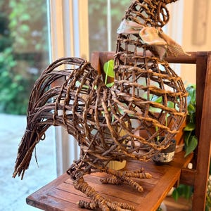 May include: A decorative rooster sculpture crafted from intertwined brown twigs and branches. The figure features a hollow, open-weave body structure, a prominent tail, and a small, light-colored plaid fabric ribbon tied around its neck.
