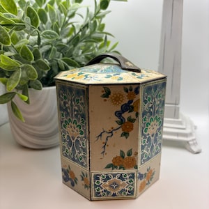 May include: A hexagonal metal canister featuring a cream-colored base with intricate floral patterns in blue, green, and yellow. The container has a matching lid with a dark metal handle on top, showcasing detailed botanical illustrations on each side.