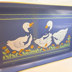 May include: A rectangular blue serving tray featuring a graphic illustration of three white geese with orange beaks and feet, each wearing a blue bow around its neck. The geese are positioned above a row of green leaves and orange flowers.