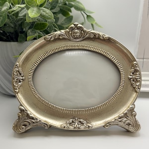 May include: An oval-shaped picture frame with a metallic champagne-colored finish and a speckled texture. The frame features ornate, raised scrollwork detailing along the edges and base, with a prominent decorative crest at the top center.