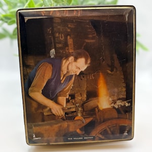 May include: A rectangular metal tin featuring a printed illustration of a blacksmith working at an anvil with a glowing forge in the background. The image depicts a person in a blue vest and brown shirt hammering metal. The text THE VILLAGE SMITHY is printed at the bottom.