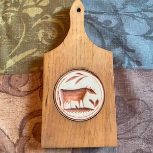 May include: A small, wooden cutting board with a handle and a circular, ceramic inset. The inset features a brown cow design on a white background. The board is made of light-colored wood and has a hole in the handle for hanging.