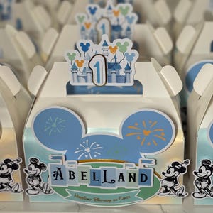 Puede incluir: Cajas blancas para recuerdos de fiesta con diseños de orejas de Mickey Mouse azules y el texto "ABELLAND". Cada caja tiene un castillo y el número "1" como adorno. Ilustraciones de Mickey Mouse están en el frente. Las cajas están decoradas con fuegos artificiales.