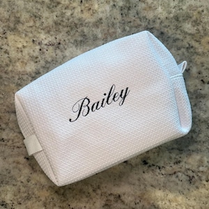 May include: White cosmetic bag with a textured waffle weave pattern. The name "Bailey" is embroidered in black script across the front. The bag has a small loop and a fabric handle.