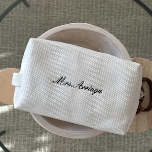 May include: A white textured cosmetic bag with the embroidered name "Mrs. Arriaga" in black script. The bag has a small loop on one side and is resting in a white bowl.