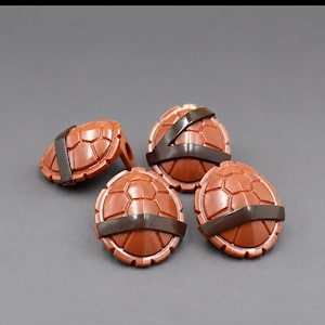 May include: Four small, round, brown plastic turtle shells, each featuring a molded hexagonal pattern and a dark brown strap detail across the center. These items are shaped like convex shields with a smooth, glossy finish and defined edges.