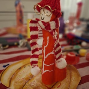 May include: A cylindrical orange wooden figurine featuring a hand-painted white branch design. The figure wears a red knitted hat with a white pom-pom, a red and white striped scarf with pom-poms, and small gold-rimmed circular glasses.