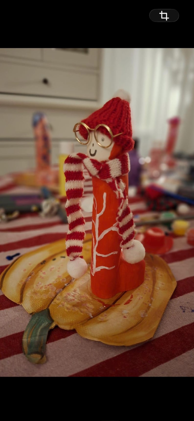 May include: A cylindrical orange wooden figurine featuring a hand-painted white branch design. The figure wears a red knitted hat with a white pom-pom, a red and white striped scarf with pom-poms, and small gold-rimmed circular glasses.