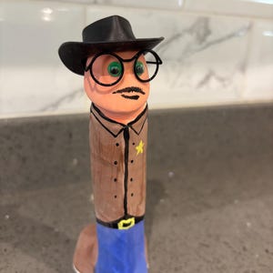 May include: A cylindrical figurine painted to resemble a sheriff, featuring a black cowboy hat, black wire-rimmed glasses, green eyes, and a mustache. The figure wears a tan shirt with a yellow star badge and blue pants with a black belt and yellow buckle.