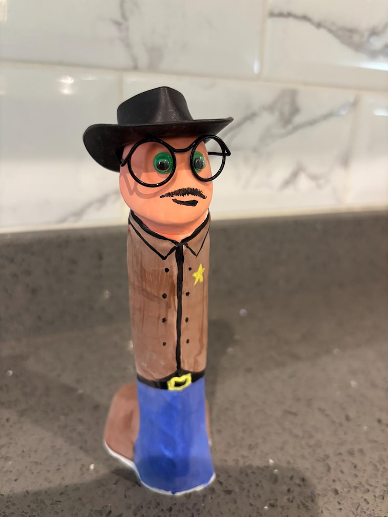 May include: A cylindrical figurine painted to resemble a sheriff, featuring a black cowboy hat, black wire-rimmed glasses, green eyes, and a mustache. The figure wears a tan shirt with a yellow star badge and blue pants with a black belt and yellow buckle.