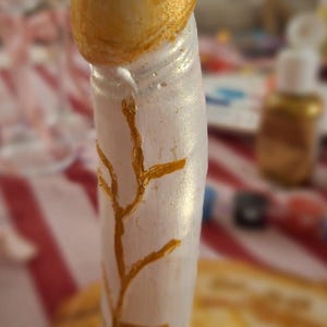 May include: A phallic-shaped sculpture featuring a metallic gold rounded top and a pearlescent white cylindrical base. The white surface is decorated with a hand-painted gold branch-like pattern that extends vertically along the side of the object.
