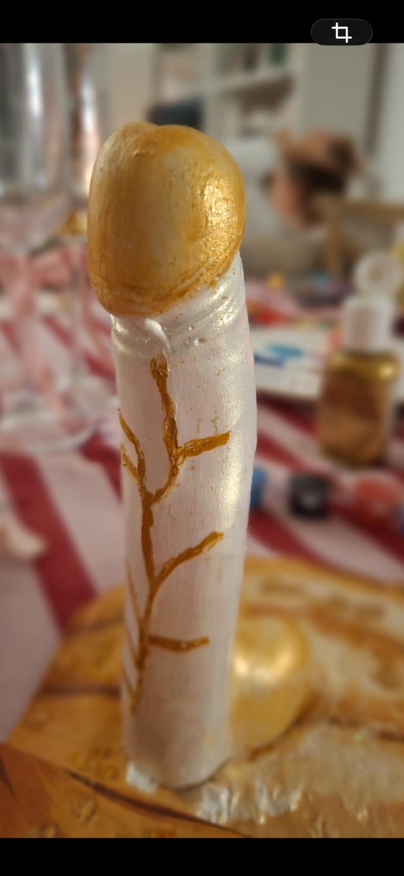 May include: A phallic-shaped sculpture featuring a metallic gold rounded top and a pearlescent white cylindrical base. The white surface is decorated with a hand-painted gold branch-like pattern that extends vertically along the side of the object.