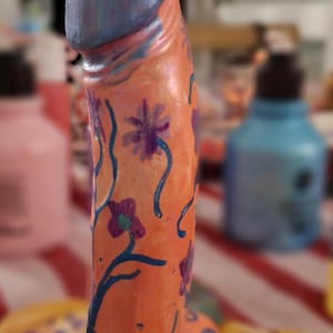 May include: A phallic-shaped sculpture painted in a light orange hue, featuring a blue-toned top section. The surface is decorated with hand-painted dark blue vines, purple flowers, and swirling patterns that extend across the entire structure.