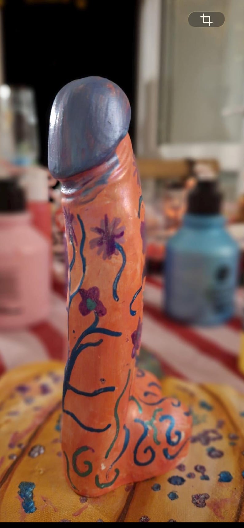 May include: A phallic-shaped sculpture painted in a light orange hue, featuring a blue-toned top section. The surface is decorated with hand-painted dark blue vines, purple flowers, and swirling patterns that extend across the entire structure.