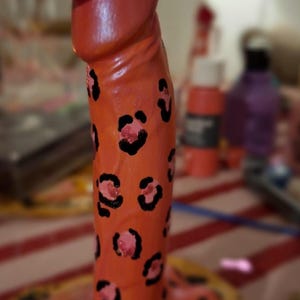 May include: A phallic-shaped sculpture painted in a bright orange hue with a black leopard print pattern throughout. The sculpture is topped with a small, pink cowboy hat featuring black spots, creating a distinct and colorful decorative object.
