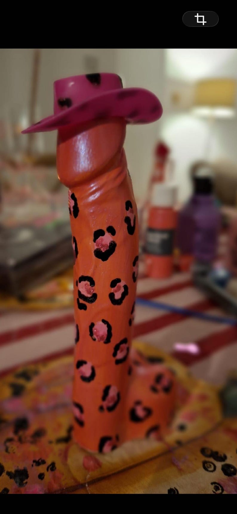 May include: A phallic-shaped sculpture painted in a bright orange hue with a black leopard print pattern throughout. The sculpture is topped with a small, pink cowboy hat featuring black spots, creating a distinct and colorful decorative object.