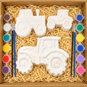 May include: A craft kit featuring three white plaster tractor figures in various sizes, accompanied by two paintbrushes and twelve small circular containers of assorted acrylic paints, all arranged within a cardboard box filled with wood shavings.