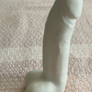 May include: A matte white phallic-shaped sculpture crafted from a solid material. The object features a smooth, rounded top section and a base with two distinct, rounded protrusions. The surface is uniform in color and texture throughout the piece.