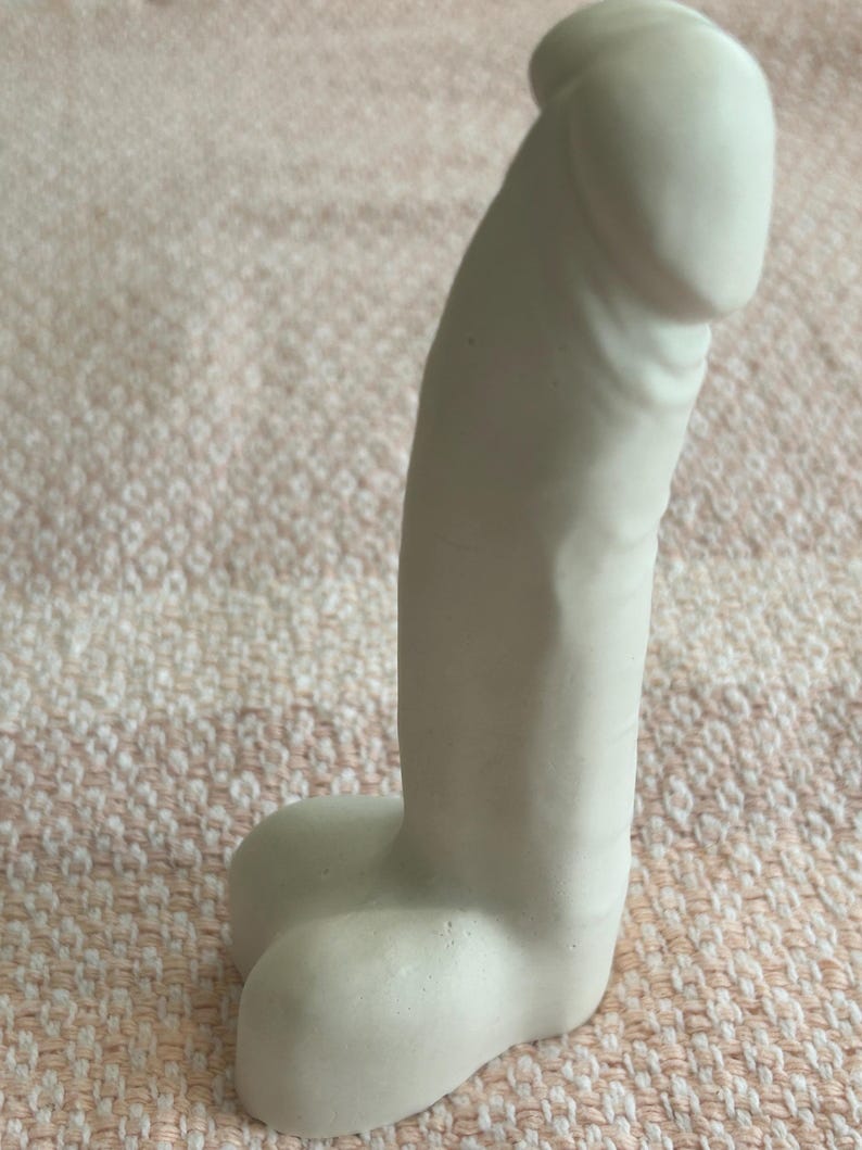 May include: A matte white phallic-shaped sculpture crafted from a solid material. The object features a smooth, rounded top section and a base with two distinct, rounded protrusions. The surface is uniform in color and texture throughout the piece.