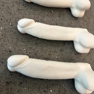 May include: Three white, phallic-shaped objects made of a smooth, matte material. Each piece features a rounded head, a cylindrical shaft, and a base with two rounded protrusions. The items are arranged in a parallel formation, showing a uniform design.