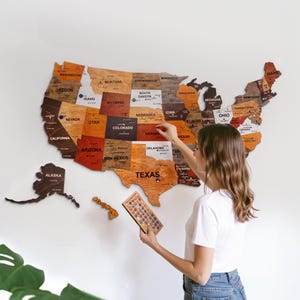 May include: A wooden United States map with individual state pieces in various wood tones. The map is mounted on a white wall. A person is holding a small wooden piece and a set of pins, presumably to mark locations.