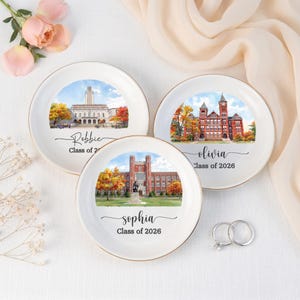 May include: Three white ceramic dishes with gold rims, each featuring a watercolor painting of a university building. The dishes are personalized with names and "Class of 2026". Two silver rings with a diamond are also present.