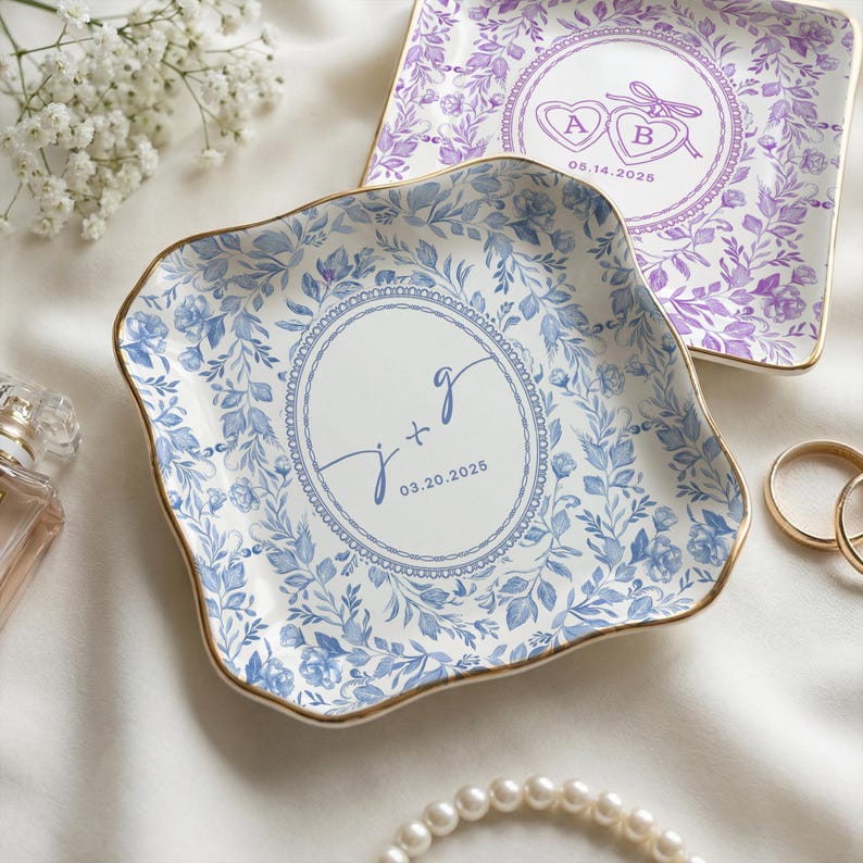 two decorative plates with floral designs, one in blue and one in purple, placed on a white surface. There are also some rings and pearls visible in the image.