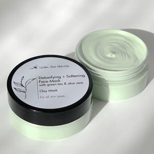 May include: Two open jars of clay face mask. One jar is closed with a black lid and a white label with text that reads "Detoxifying + Softening Face Mask with green tea & aloe vera Clay Mask For all skin types". The mask is light green.