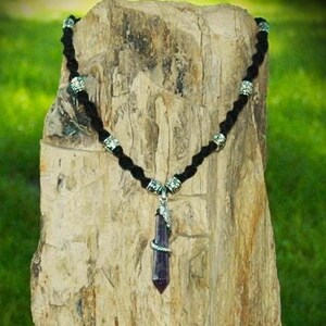 May include: A black cord necklace with silver-toned beads and a pendant. The pendant features a dark purple crystal wrapped with a silver-toned metal design. The necklace is displayed against a textured, light brown background with a green backdrop.