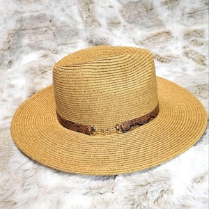 Op de afbeelding: Een beige strooien fedora-hoed met een brede rand en een gestructureerde kroon. De hoed is versierd met een bruine band met slangenleerpatroon rond de basis van de kroon, afgewerkt met een goudkleurig metalen bitdetail aan de voorzijde.