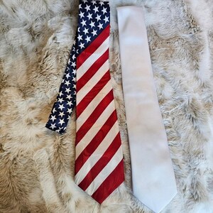 May include: Two neckties displayed side by side. One tie features a classic American flag pattern with a blue field of white stars at the top and diagonal red and white stripes below. The second tie is a solid, plain white color with a smooth finish.