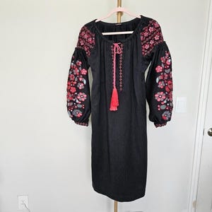 以下が含まれることがあります： 袖と胴に花の刺繍が施された黒いドレス。ドレスは、ピンクのタッセルとドローストリングが付いた丸いネックラインです。袖は長くふっくらとしており、ドレスは金のハンガーに掛けられています。