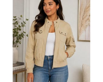May include: A beige utility-style bomber jacket featuring a stand-up collar, a full-length front zipper, two chest pockets with buttoned flaps, and ribbed cuffs at the sleeves and hem. The jacket is worn over a plain white top and blue denim jeans.