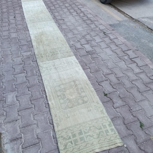May include: A long, narrow runner rug featuring a faded, light green and beige color palette. The rug displays a repeating geometric pattern with central medallion motifs and intricate border designs, showcasing a weathered, low-pile texture.