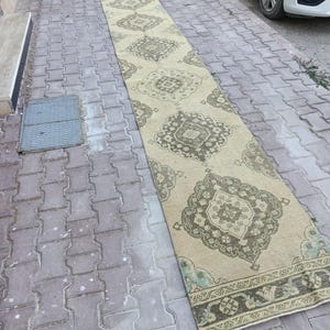 May include: A long, narrow runner rug featuring a light beige base colour with a repeating pattern of dark olive green geometric medallions. The rug has a decorative border with intricate floral and geometric motifs in matching olive green tones.