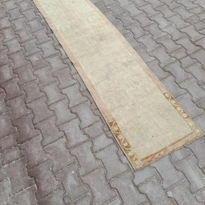 Può includere: Un lungo tappeto rettangolare dall'aspetto sbiadito e vintage. Il tappeto presenta un campo centrale beige con un motivo sottile e un bordo marrone. È posizionato su una superficie di mattoni grigi.