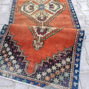 May include: An orange and blue vintage runner rug with geometric patterns. The rug features a central diamond design with cream, blue, and purple accents. The border is a dark blue with a repeating floral pattern. The rug is folded over.